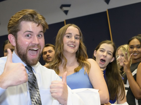 A white male medical student with a beard smiles and gives two thumbs-up as several female medical students smile behind him. 