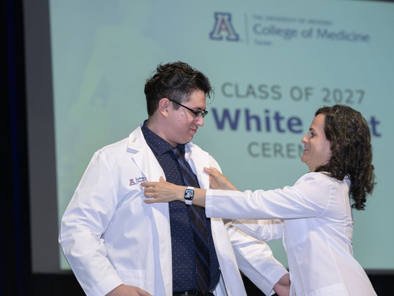 A male medical student stands on stage as a female faculty member helps him put on his white medical coat. 