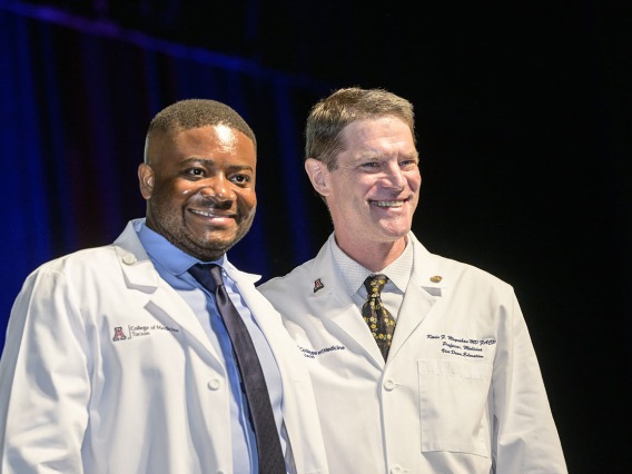 A young black medical student stands next to a white male faculty member. Both are smiling and wearing white medical coats. 