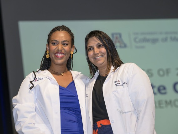 A dark-skinned femal medical studeent stands next to a femal faculty member. Both are wearing white medical coats and smiling. 