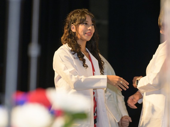 A Hispanic female medical student in a white coat smiles as she reaches out to shake hands with a faculty member. 