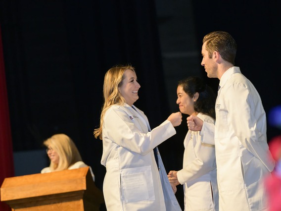 A light-skinned female medical student with long blonde hair smiles as she fist-bumps a male faculty member after receiving her white coat. 