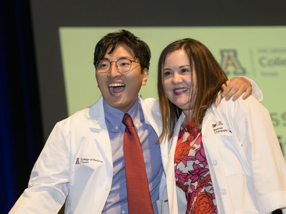 A young Asian male medical student stands next to a female professor with his arm on her shoulder. Both are smiling and wearing white medical coats. 