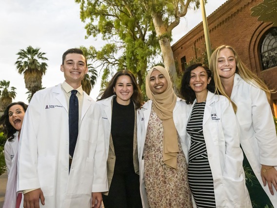 A female medical school professor making a silly face peeks around a group of five people in white medical coats as they pose for a photo. 