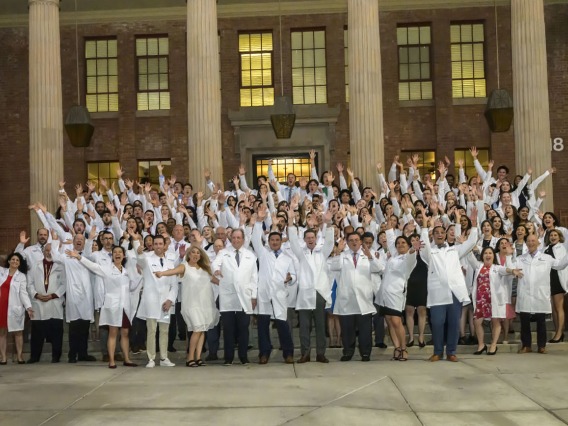 A group of over 100 medical school students and faculty wearing white coats raise their hands cheering outside a building with large pillars. 