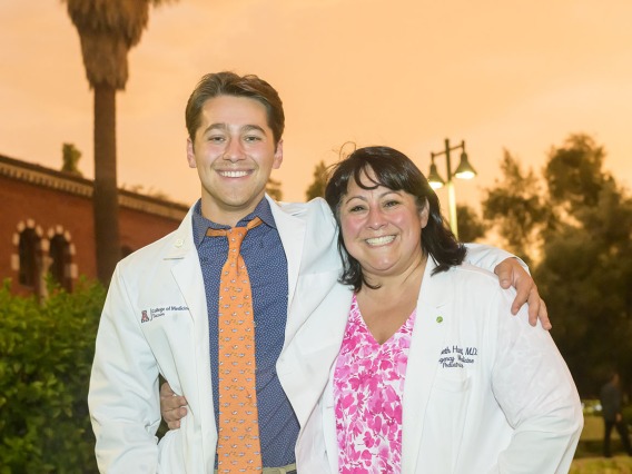 A young white male medical student stands with his arm around his mom, both are wearing white medical coats and smiling. 
