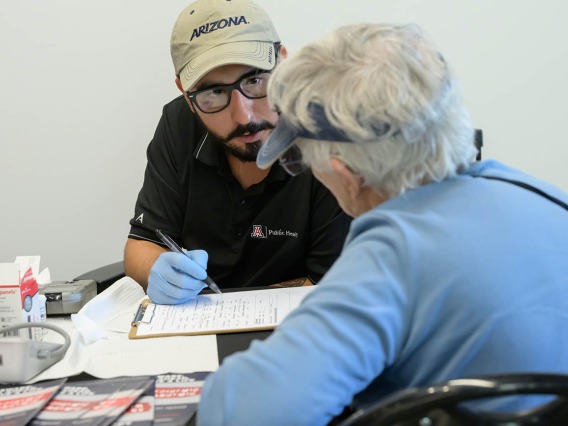 A man with a dark goatee, glasses and a baseball cap wearing medical gloves fills out paperwork for a woman with short gray hair. 