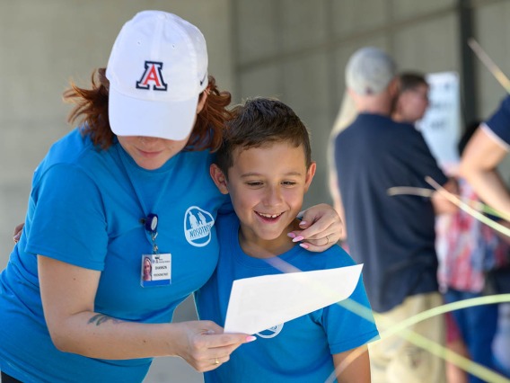 A woman with a white Arizona baseball hat and bright blue shirt has her arm around her son while showing him a piece of paper. 