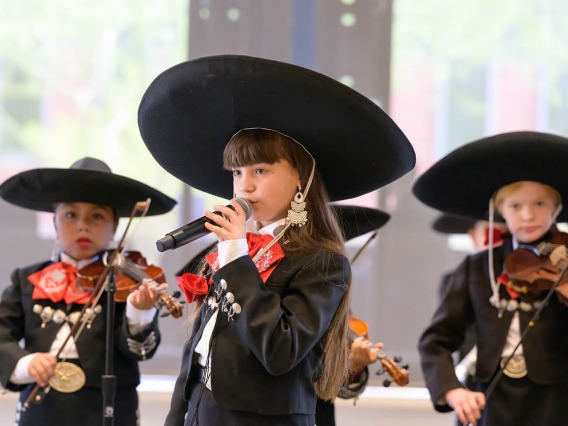 A young girl wearing a large black hat and black mariachi suit sings into a microphone as other kids dressed the same play violins in the background. 