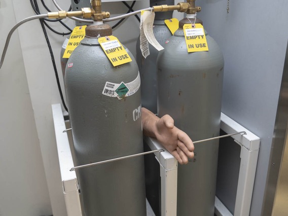 A hand sticks out from between two larg metal tanks with warning labels on them in a lab setting. 