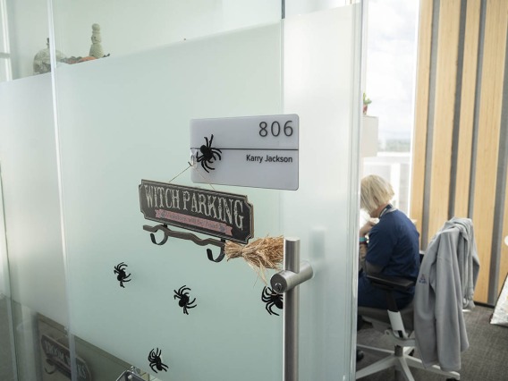 View looking through an open glass office door with a sign that reads, "Witch Parking" with a small witch's broom hanging from it. 