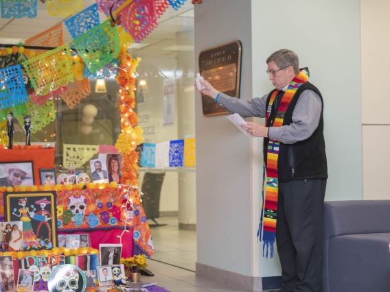 Chaplain Joe Fitzgerald stands beside the colorful Dia de los Muertos altar with his hand raised as he reads a blessing. 