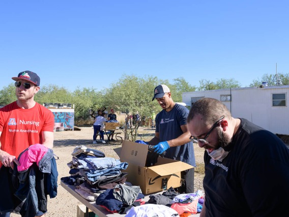 Three young men, all Doctor of Nursing Practice residents, sort donated clothes at a table covered in clothing in a dirt lot. 