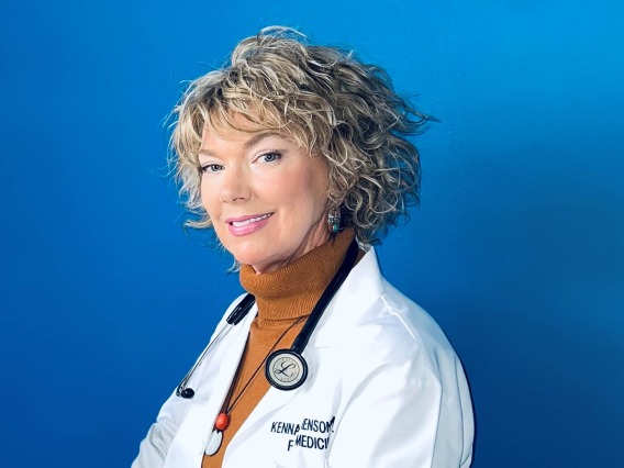 Woman doctor with short, curly blonde hair stands against a blue background, wearing a burnt orange turtleneck, white coat with her name on it and a stethoscope. 