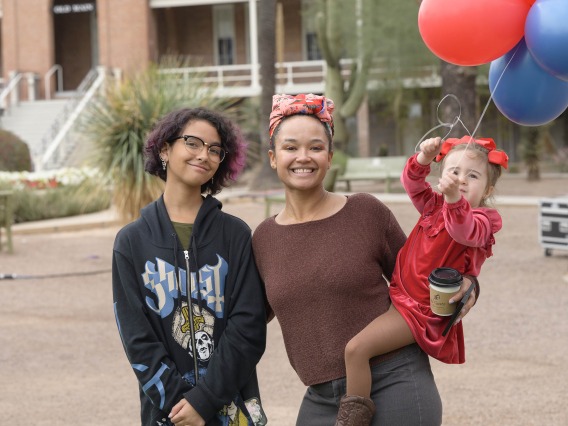 Two women stand next to each other, smiling. One is holding a small child who is holding balloons.