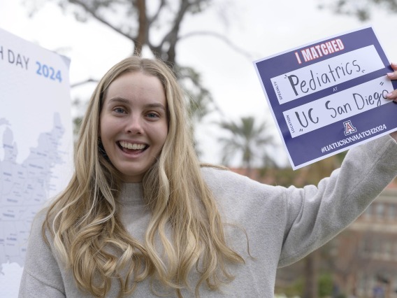 A smiling female, fourth-year medical student from the University of Arizona College of Medicine – Tucson holds a sign that says “I Matched” as she stands next to a map of the United States.