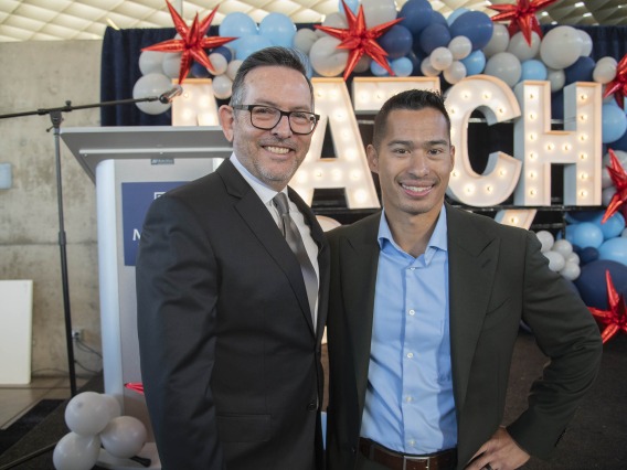 Two men in suitcoats smile as they stand in front of a stage with balloons at the University of Arizona College of Medicine – Phoenix Match Day event.