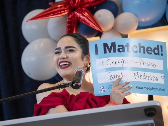 A smiling female, fourth-year medical student from the University of Arizona College of Medicine – Phoenix stands at a podium holding up a sign that says “I Matched” as she announces where she will be conducting her residency.