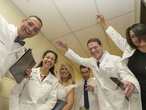 Six medical school faculty members wearing white coats laugh and make funny gestures with their arms while standing in a hall. 