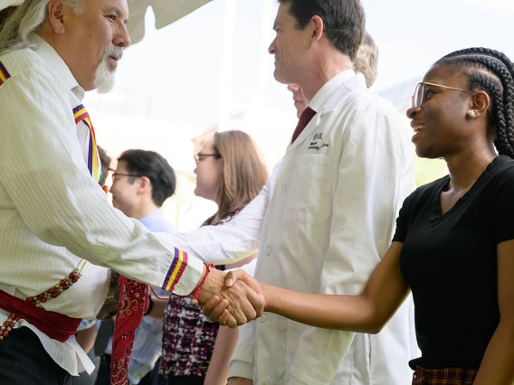 An older man in a Native American-style shirt shakes the hand of a young woman. 