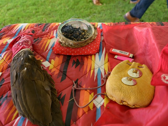Table with red tablecloth and an eagle feather fan, an abalone shell a leather pouch sitting on it. 