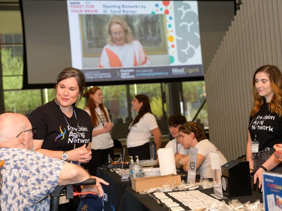 Two women wearing t-shirts that read "Precision Aging Network" talk with an older man in a wheelchair inside a large open room with other people in the background. 