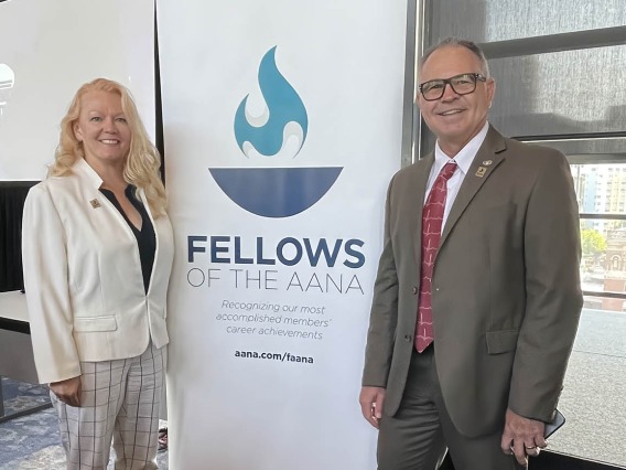 A woman with long blonde hair and a white suite coat stands with a man in a suite on either side of a banner that reads AANA Fellows.