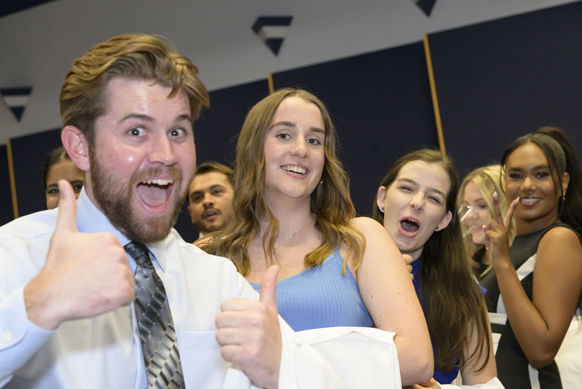 A white male medical student with a beard smiles and gives two thumbs-up as several female medical students smile behind him. 