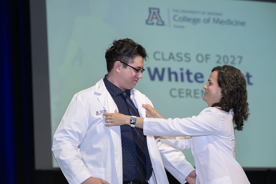 A male medical student stands on stage as a female faculty member helps him put on his white medical coat. 