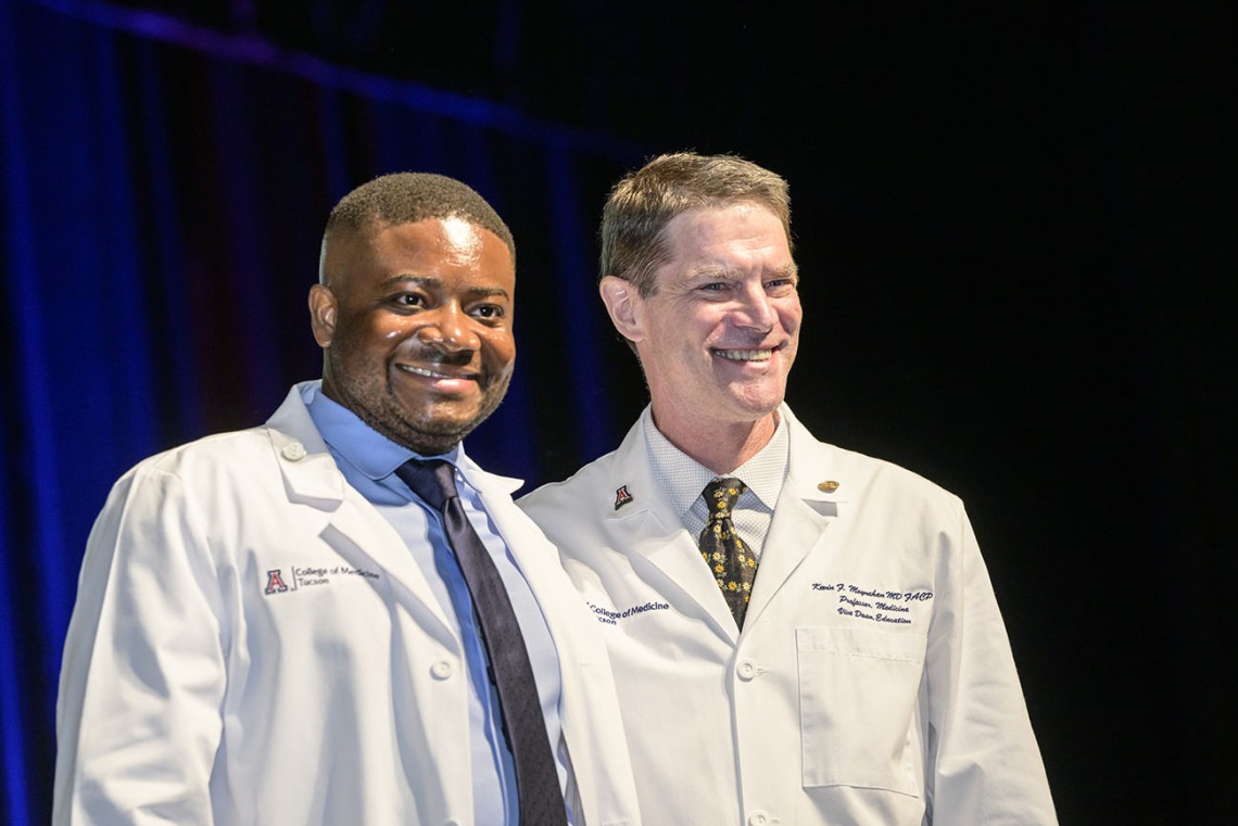 A young black medical student stands next to a white male faculty member. Both are smiling and wearing white medical coats. 