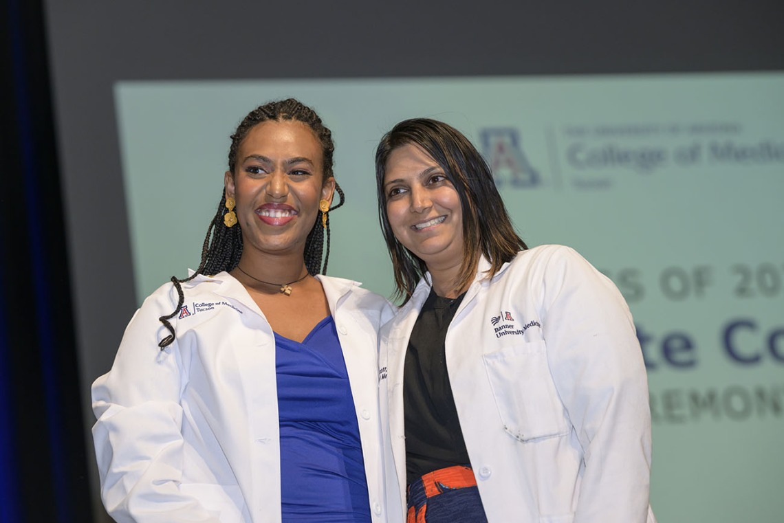 A dark-skinned femal medical studeent stands next to a femal faculty member. Both are wearing white medical coats and smiling. 