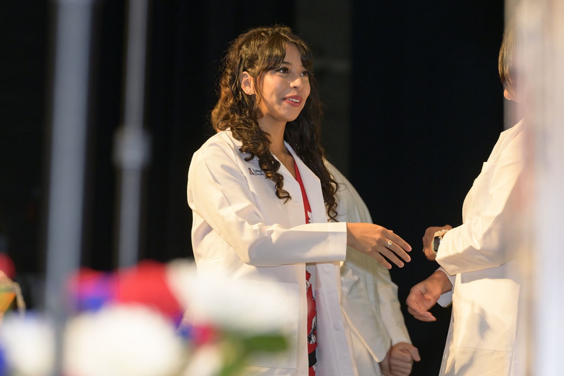 A Hispanic female medical student in a white coat smiles as she reaches out to shake hands with a faculty member. 