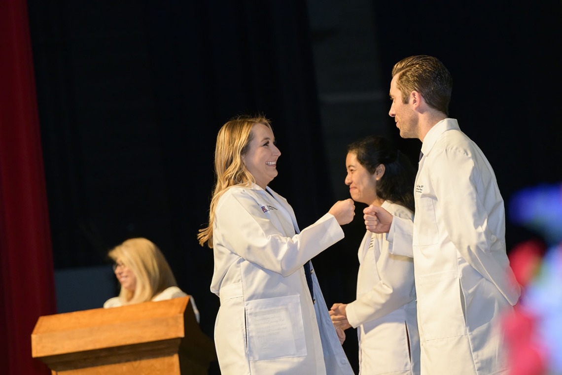 A light-skinned female medical student with long blonde hair smiles as she fist-bumps a male faculty member after receiving her white coat. 