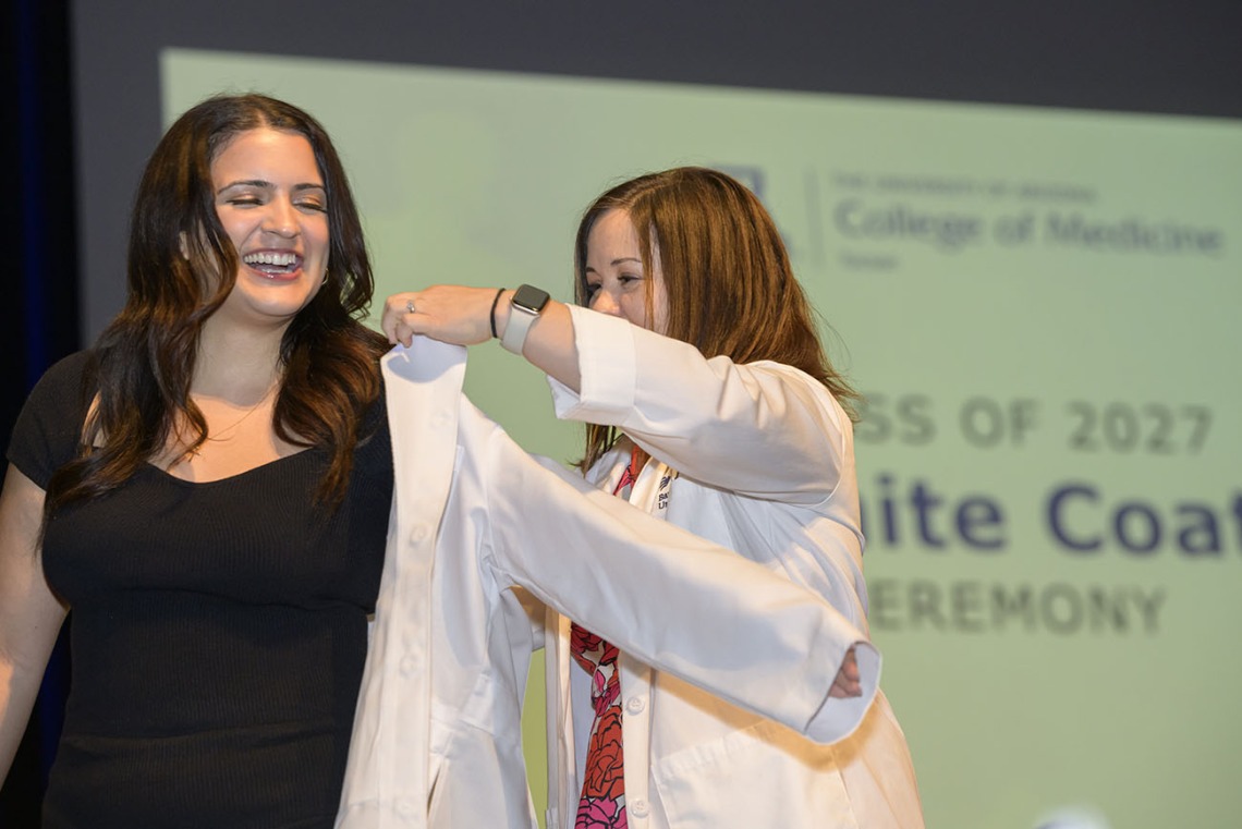 A young female medical student with long dark hair smiles as a female faculty member helps her put on her white coat. 
