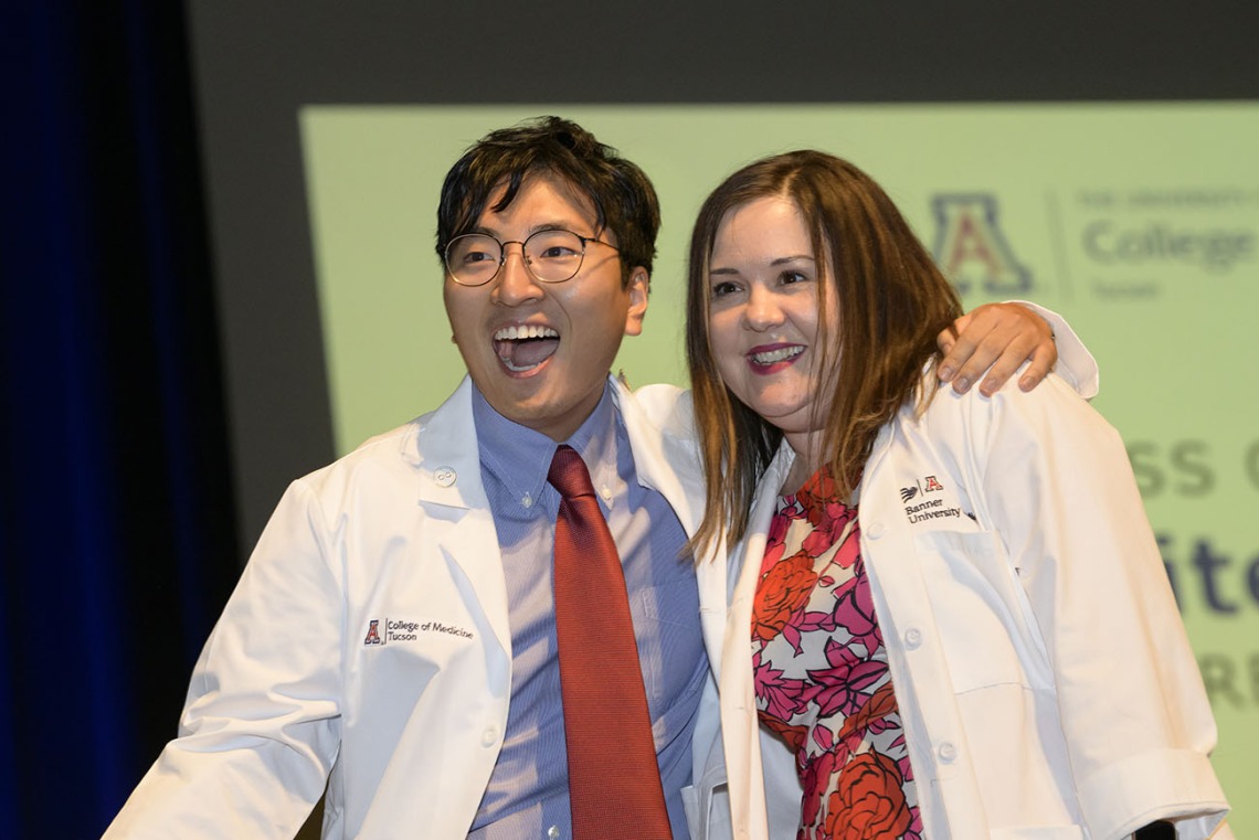 A young Asian male medical student stands next to a female professor with his arm on her shoulder. Both are smiling and wearing white medical coats. 