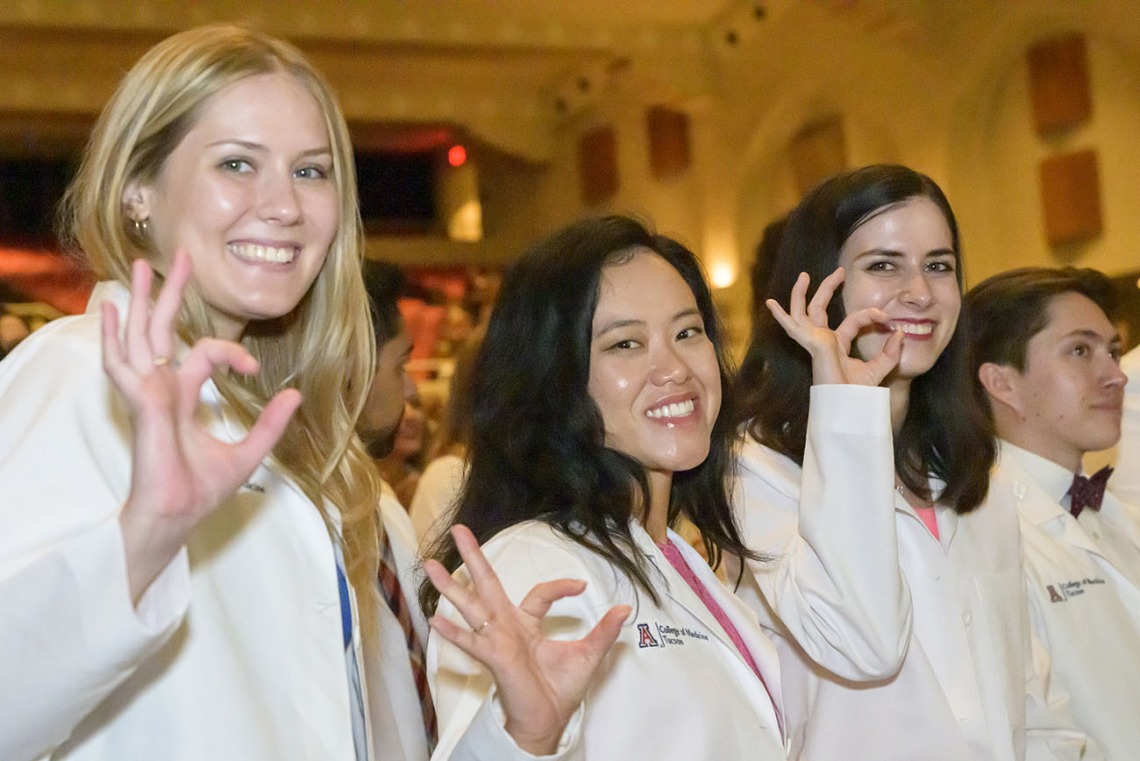 Three female medical students in white coats smile while holding up their hands make the Arizona Wildcat sign.