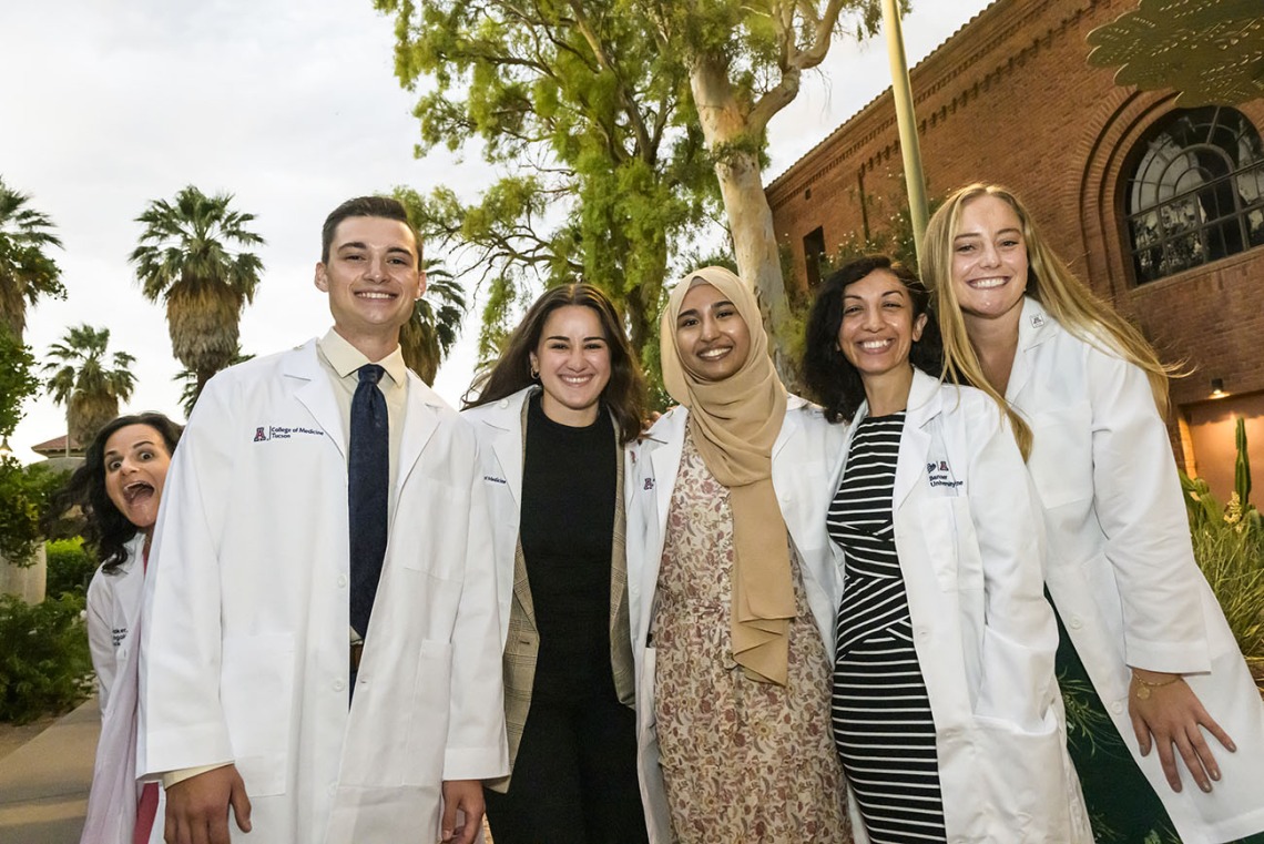A female medical school professor making a silly face peeks around a group of five people in white medical coats as they pose for a photo. 