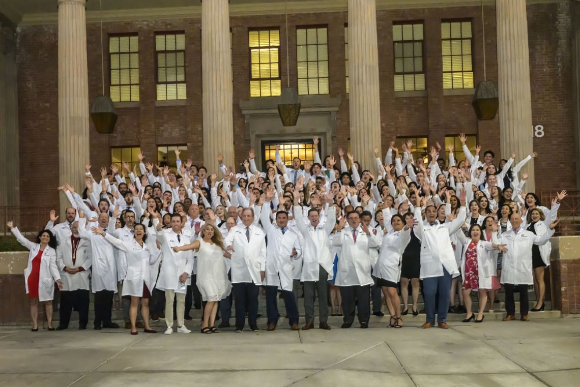 A group of over 100 medical school students and faculty wearing white coats raise their hands cheering outside a building with large pillars. 