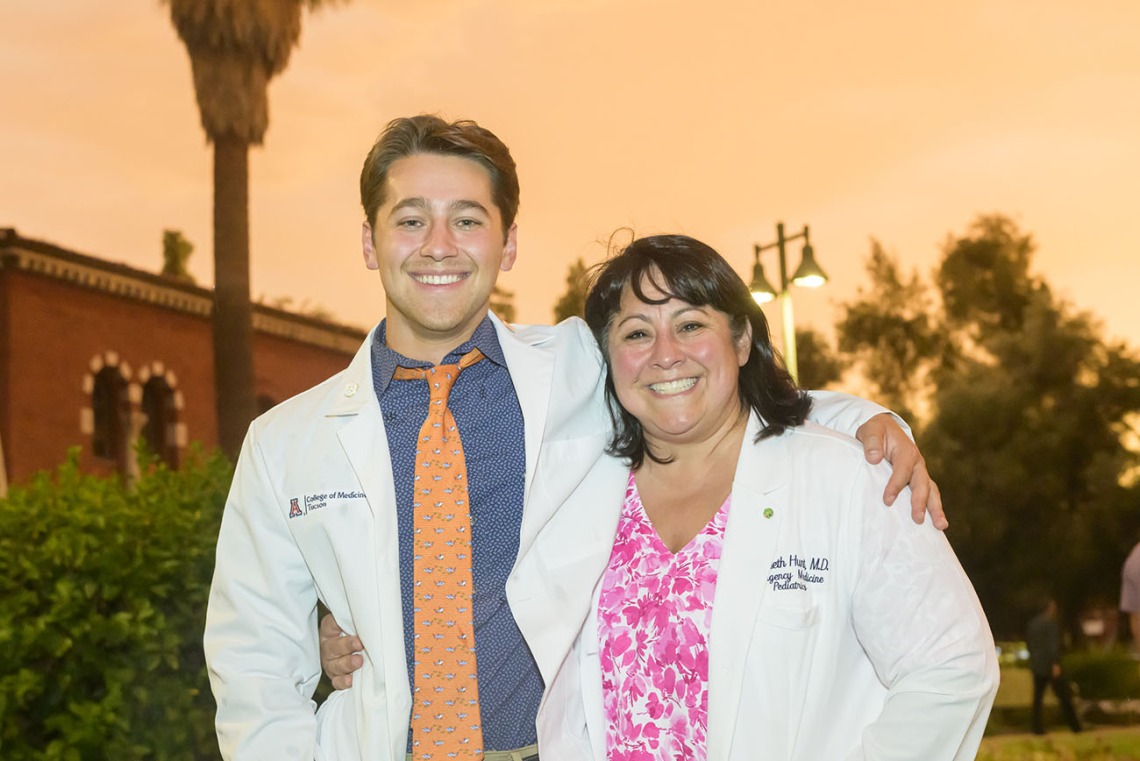 A young white male medical student stands with his arm around his mom, both are wearing white medical coats and smiling. 
