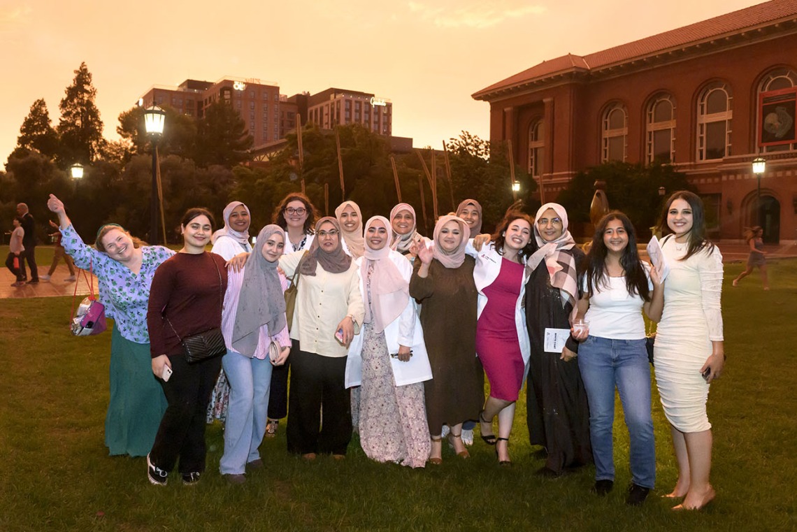 A group of about 15 women stand together, many of them medical students wearing white coats, smiling outside in the grass. 