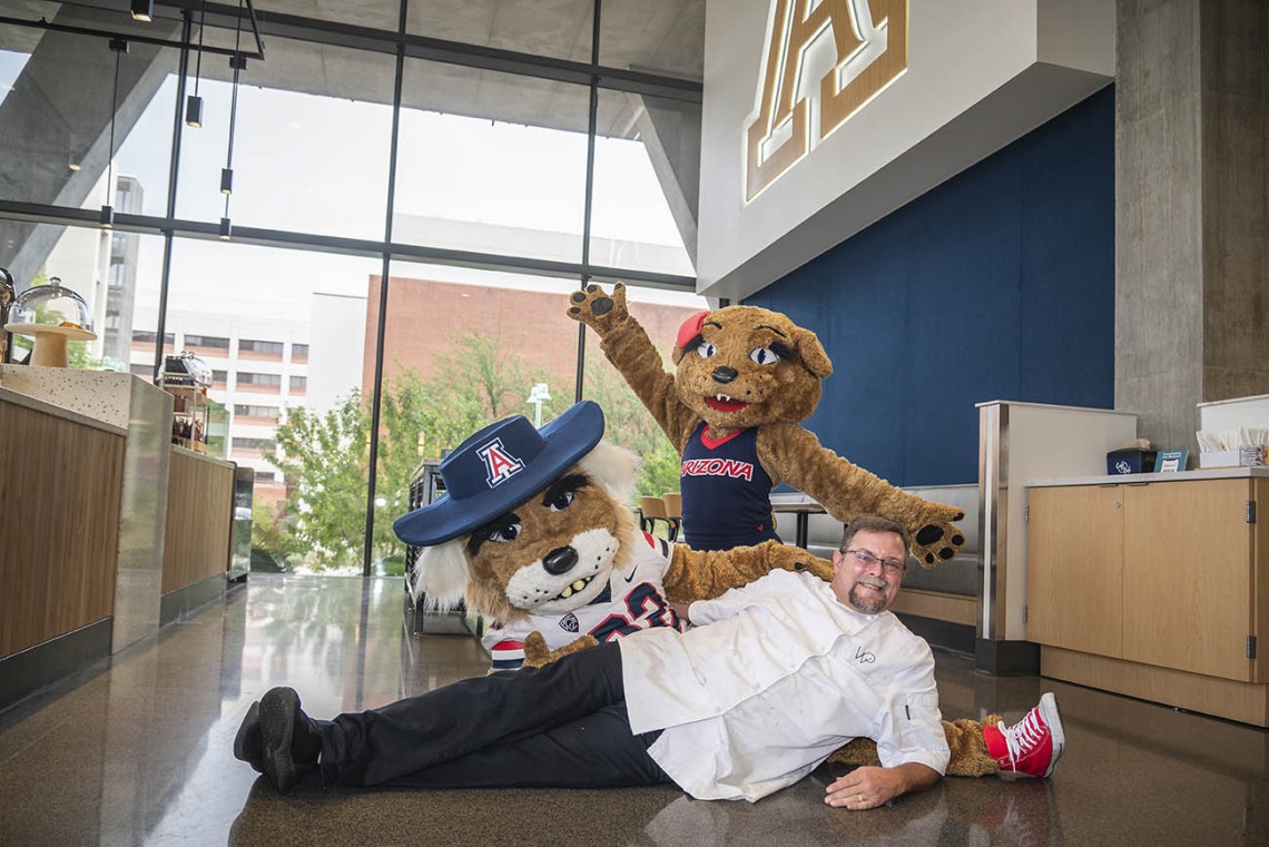 A male chef lays on his side next to the Wilbur the Wildcat mascot as the Wilma Wildcat mascot stands behind them with arms raised. 