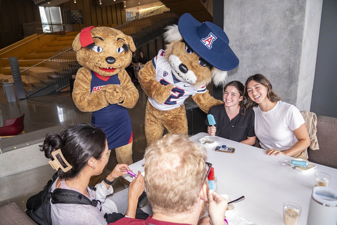 Wilma and Wilbur Wildcat mascots visit with young adults at a table while they eat. 