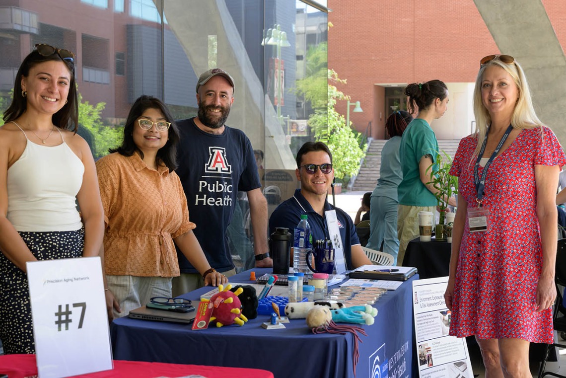 Five employees from the Zuckerman College of Public Health stand behind information tables in an outside setting. 