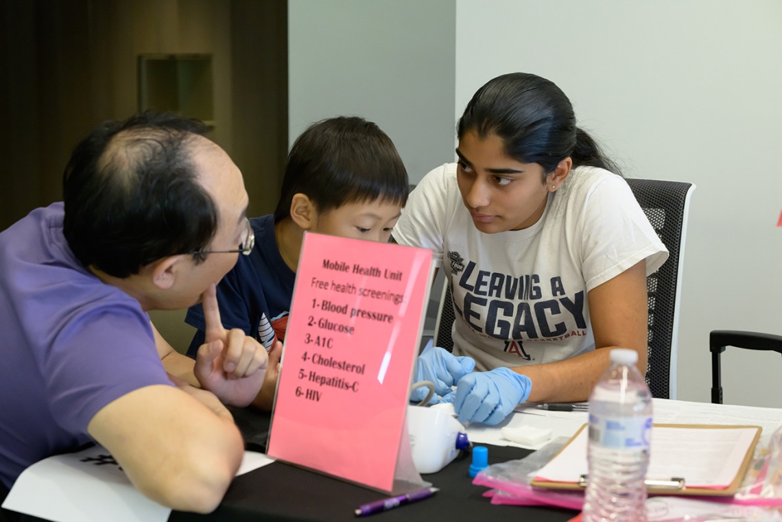 A young woman sits at a table wearing medical gloves and talks with a father and son at the Mobile Health Unit information table. 