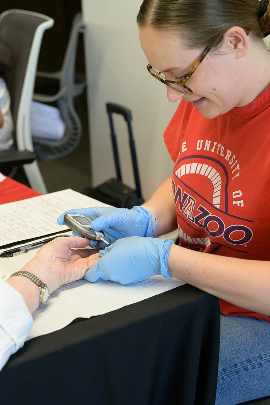 A woman in a red University of Arizona t-shirt wearing medical gloves takes a blood sample from the finger of someone's hand.