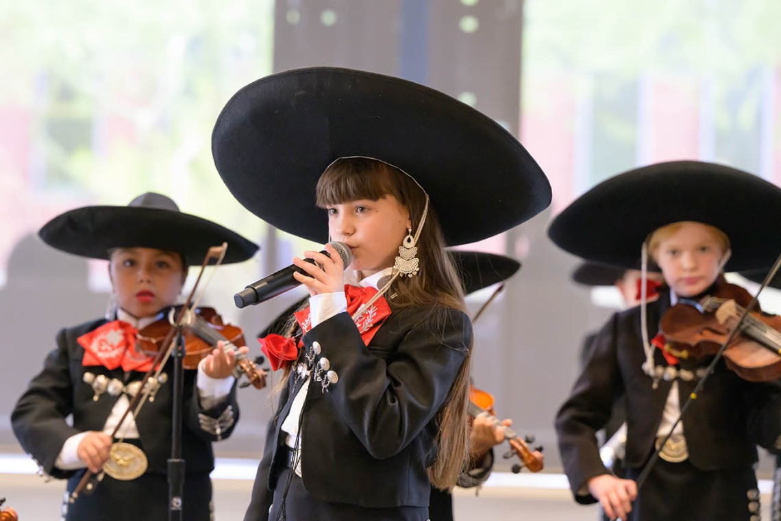 A young girl wearing a large black hat and black mariachi suit sings into a microphone as other kids dressed the same play violins in the background. 