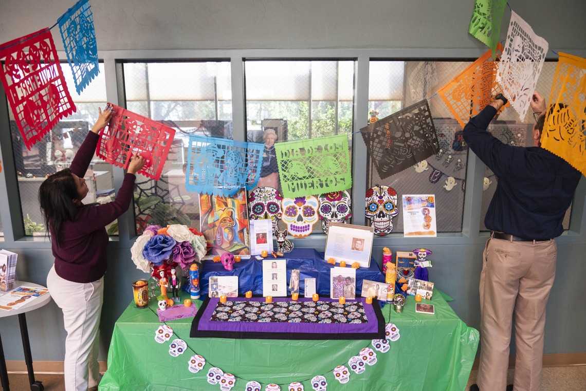 Two people hang tissue paper decorations over a brightly decorated Dia de los Muertos altar with skull decorations and photographs. 