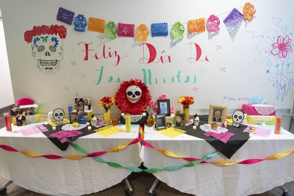 A long table is set up as a Dia de los Muertos altar with colorful streamers and skull decorations. There are photos and flowers on the table. 