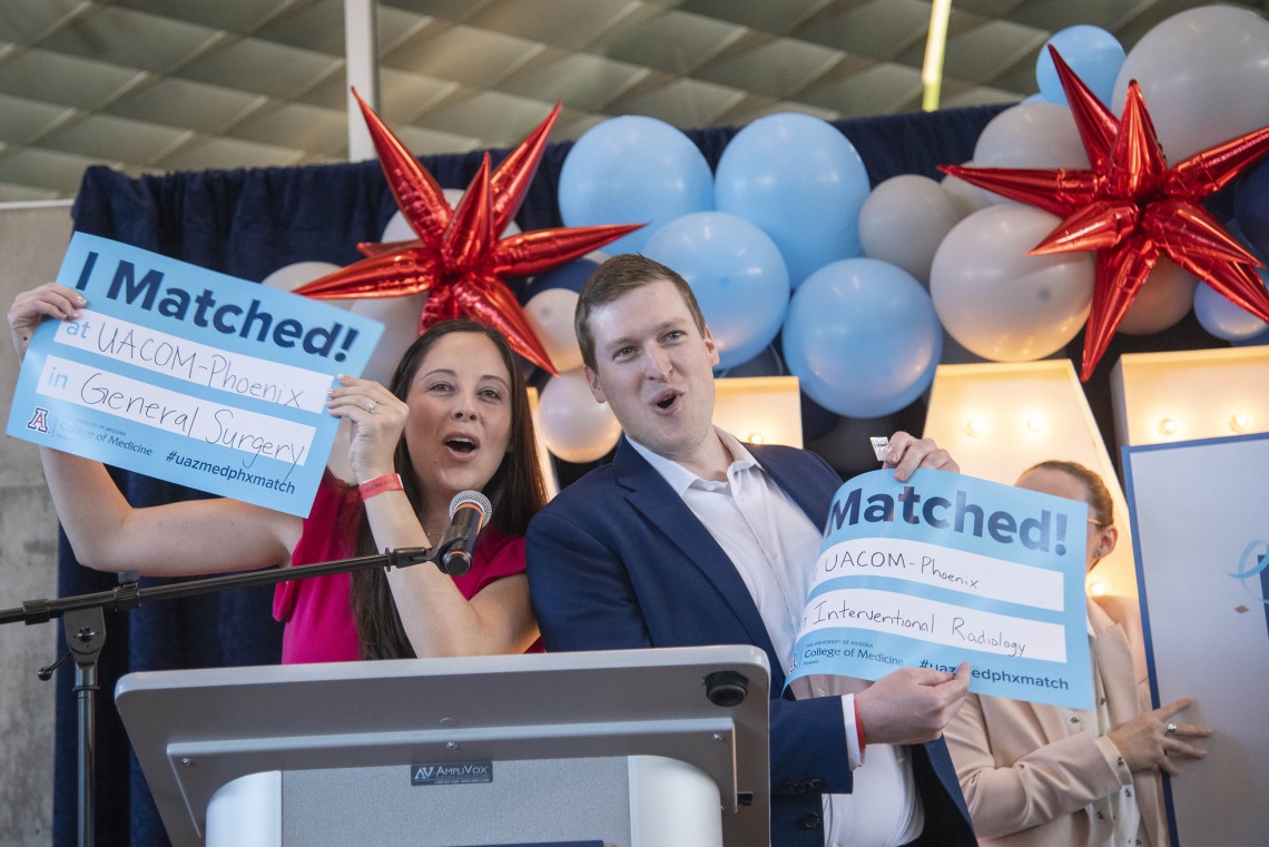 Two fourth-year medical students from the University of Arizona College of Medicine – Phoenix stand at a podium holding up signs that say “I Matched!” as they announce where they are headed for residency. 