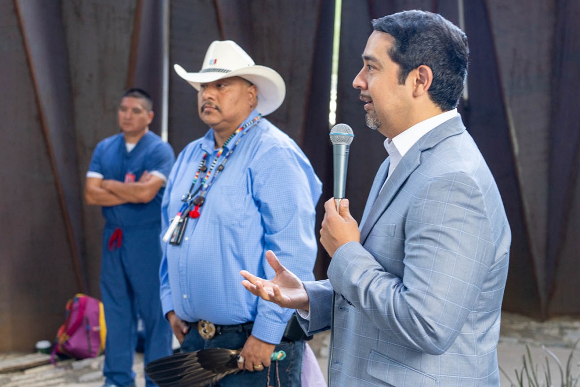 A man in a gray suite holding a microphone talks in an outdoor setting with a man in a straw cowboy had and beeded necklace stands in the background. 